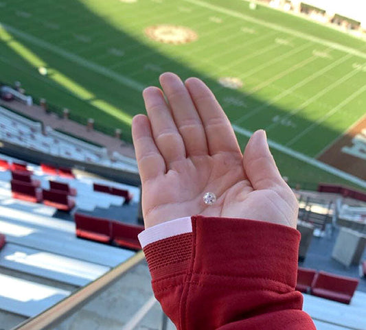 Close-up of a hand holding a sparkling diamond ring, with the empty football stadium field in the background. The person is wearing a red jacket, and the image captures the excitement of a special moment in a unique setting.