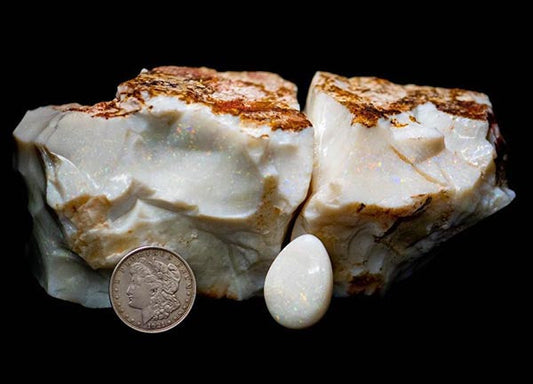 Close-up of two rough opal rocks and a polished opal stone, with a coin placed next to them for scale. The opals are white with a hint of colorful iridescence, set against a black background.