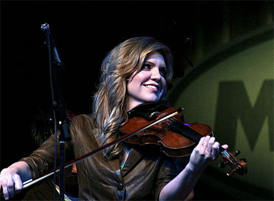 Smiling woman playing the violin on stage, wearing a brown jacket with a spotlight shining on her as she performs.