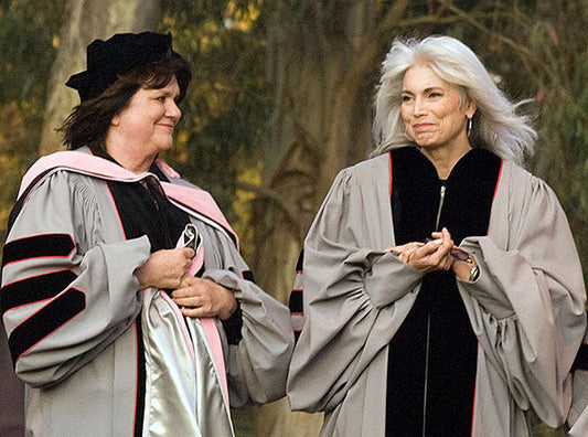 Two women in academic regalia at a graduation ceremony, smiling and engaging in conversation, one in a black and pink doctoral gown and the other in a gray and black doctoral gown, celebrating their academic achievement.