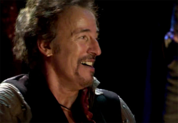 Legendary musician Bruce Springsteen smiling on stage during a live performance, captured in a candid moment. The image highlights his joyful expression and distinctive curly hair.