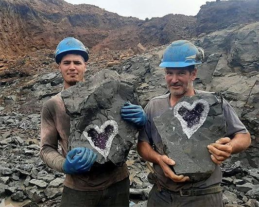 Two miners holding large rocks with purple heart-shaped crystals in a quarry, wearing blue helmets and gloves.