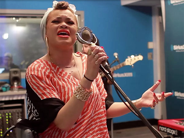 Female singer passionately performing into a vintage microphone in a recording studio, wearing a patterned orange blouse and gold jewelry, with a band in the background.