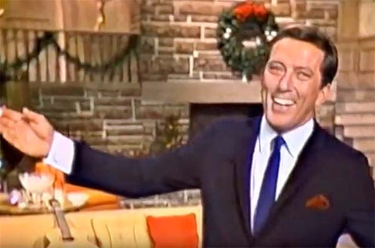 Andy Williams smiling and gesturing while hosting his Christmas special show, with a fireplace and festive decorations in the background.