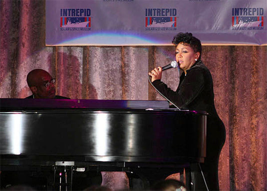 Singer performing on stage at the Intrepid Museum, with a pianist accompanying her on a grand piano during a live performance event.