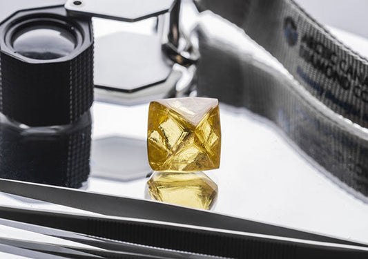 Close-up image of a yellow rough diamond placed on a reflective surface with a loupe and tweezers nearby, used for diamond inspection.