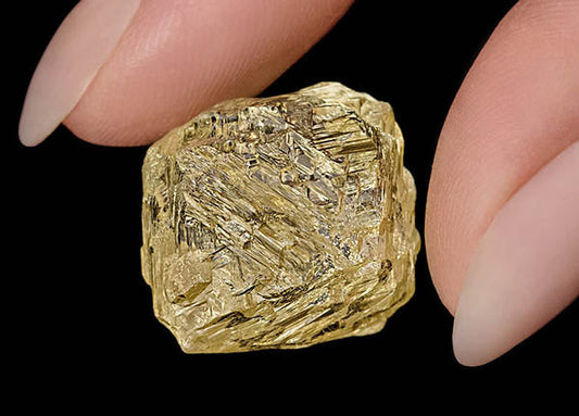 Close-up of a hand holding a rough, uncut yellow diamond with visible natural crystal formations. The diamond is set against a black background, showcasing its unique texture and shape.