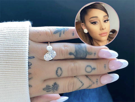 A close-up of Ariana Grande's hand showcasing her engagement ring with a diamond and pearl, featuring visible tattoos and manicured nails. An inset image of Ariana Grande's face is included in the corner.