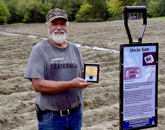 Man proudly holding an award in front of an informational sign about Uncle Sam gold mine, with a field in the background, showcasing his achievement as a certified 'Craterhead.