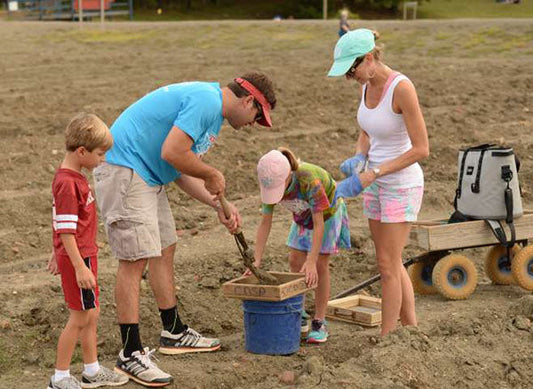 A family of four, including two children, working together to dig and collect items from the soil at an outdoor excavation site, with a cart nearby for carrying the items.
