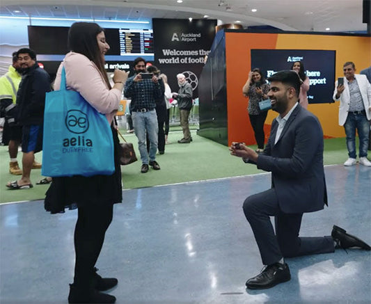 Man proposing marriage to woman at Auckland Airport, with onlookers taking photos in the background.
