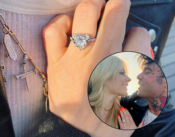 Close-up of a woman's hand showing an engagement ring with a large heart-shaped diamond, with a photo of the couple smiling at each other in the background.