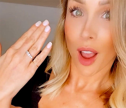 Smiling woman showing off her engagement ring, with her hand raised and fingers spread. The ring features a beautiful design with multiple sparkling stones. She has light blonde hair and is wearing natural makeup, with an excited expression on her face.