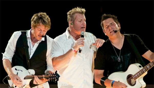 Rascal Flatts band members performing on stage, with one guitarist playing an electric mandolin, another guitarist holding an acoustic guitar, and the lead vocalist singing into a microphone.