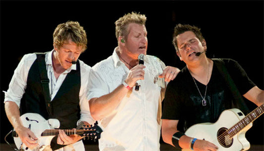 Rascal Flatts band members performing on stage, with one guitarist playing an electric mandolin, another guitarist holding an acoustic guitar, and the lead vocalist singing into a microphone.