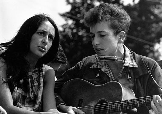 Black and white photo of Joan Baez and Bob Dylan performing together with Joan singing and Bob playing guitar and harmonica.
