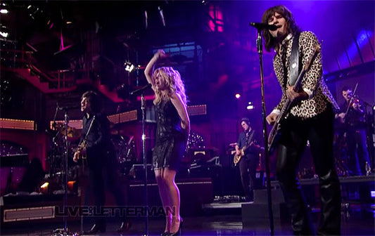 Rock band performing live on stage during a television show, with a singer and musicians playing guitars under colorful stage lighting.