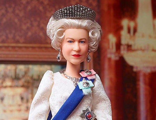 Close-up of a collectible Queen Elizabeth II doll, dressed in a royal gown with a blue sash, crown, and jewelry. The doll is styled with white curly hair and earrings, showcasing intricate details of the royal attire.
