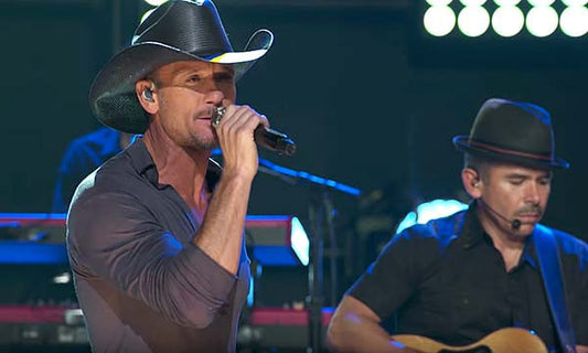 Tim McGraw performing on stage during a concert, holding a microphone and singing, with a fellow musician playing guitar in the background.