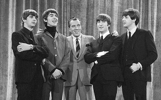 The Beatles posing with Ed Sullivan on his show, with Ringo Starr, George Harrison, John Lennon, and Paul McCartney in black and white 1960s attire.