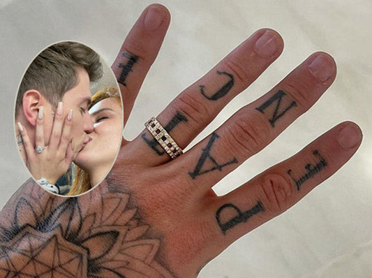 Close-up of a hand with a diamond wedding band and tattoos, showing the engagement ring on the ring finger, with an inset photo of the couple sharing a kiss.