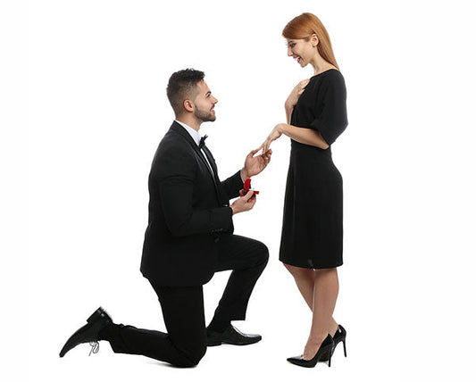A man kneeling with an engagement ring in a box, proposing to a woman who is smiling and looking at the ring, in formal attire.