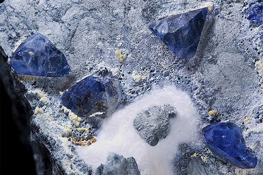 Close-up of rough blue sapphires embedded in stone, showcasing their natural, uncut form. The sapphires are surrounded by white mineral deposits and rocky textures, highlighting their raw beauty.