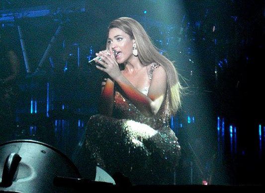 Pop singer performing on stage in a sparkling dress with long hair, singing into a microphone under stage lights.
