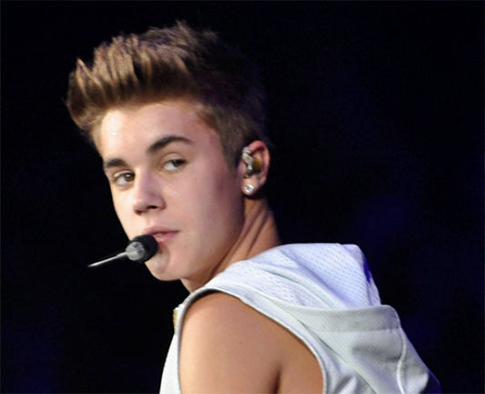 Close-up image of Justin Bieber performing on stage, wearing a sleeveless hoodie and microphone, with a confident look over his shoulder.