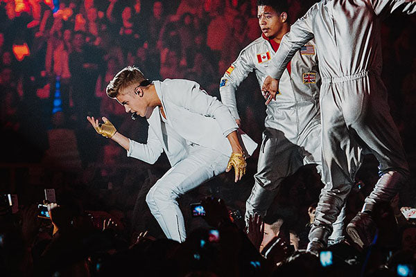 Justin Bieber performing on stage during a concert, wearing a white suit and gold gloves, with dancers in silver outfits and a lively crowd in the background.