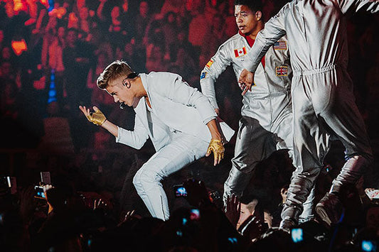 Justin Bieber performing on stage during a concert, wearing a white suit and gold gloves, with dancers in silver outfits and a lively crowd in the background.