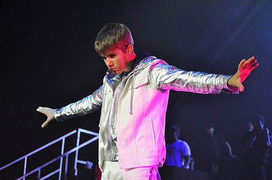 Justin Bieber performing on stage during a live concert, wearing a silver jacket and white pants, with arms extended. The image captures him in a dramatic moment with vibrant stage lighting in the background.