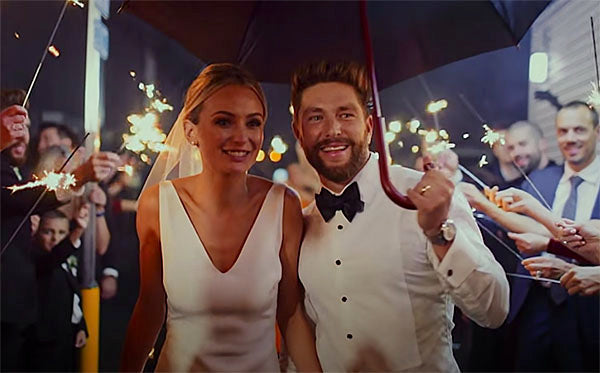Newlywed couple smiling while walking under an umbrella with sparklers held by guests in the background, celebrating their wedding day.