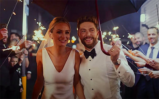 Newlywed couple smiling while walking under an umbrella with sparklers held by guests in the background, celebrating their wedding day.