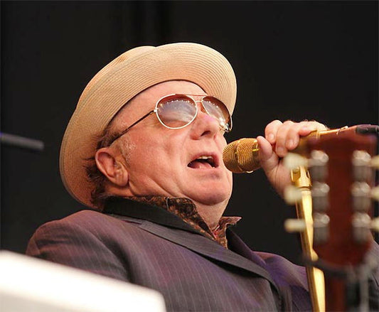 Legendary musician performing on stage, wearing a beige fedora hat, sunglasses, and a patterned shirt. He is singing into a golden microphone with a guitar in the background, showcasing his iconic performance style.