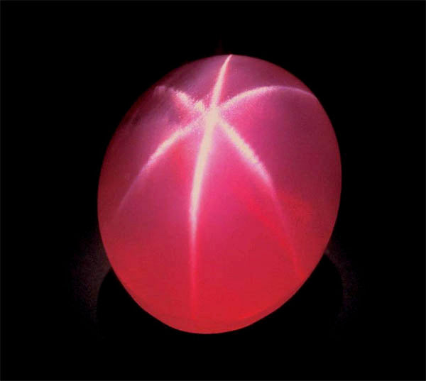 Close-up of a glowing pink gemstone with a star-like reflection on a dark background, showcasing its unique light refraction.