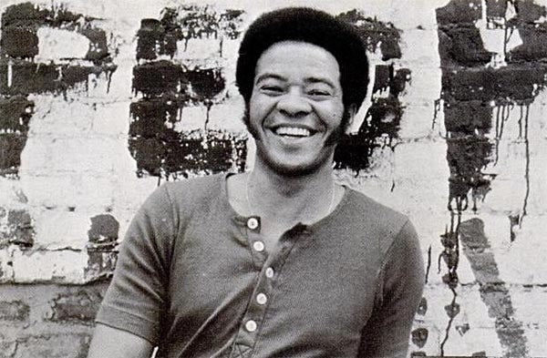 Black and white photo of smiling Bill Withers, wearing a casual shirt, standing in front of a brick wall with graffiti.