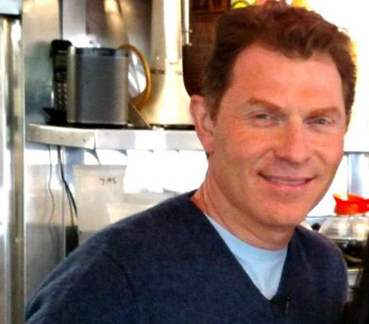 Celebrity chef Bobby Flay smiling in a kitchen setting, wearing a blue sweater and standing near kitchen equipment.