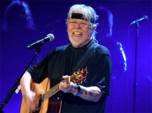 Musician Bob Seger performing on stage with a guitar, smiling and wearing a black headband during a live concert.