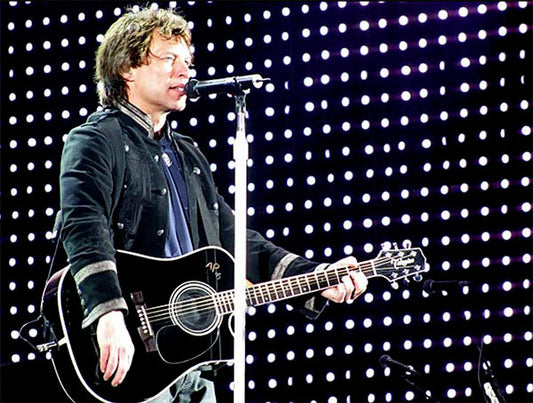 Jon Bon Jovi performing live on stage with an acoustic guitar, singing into a microphone, with bright LED lights in the background.