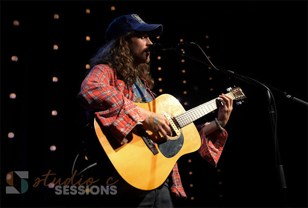A musician performing on stage with an acoustic guitar, wearing a plaid shirt and a cap, in front of a backdrop of lights during a live performance.