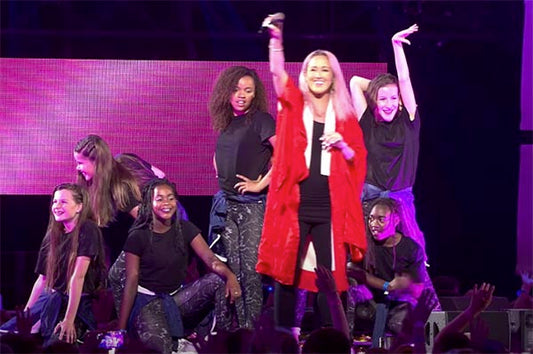 Pop singer performing on stage with a group of dancers during a live concert, wearing a red jacket and surrounded by young dancers in black t-shirts and camo pants.