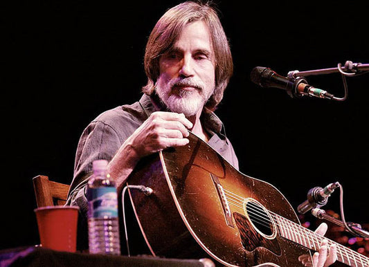 Jackson Browne performing live on stage with his guitar. The musician, known for his classic rock and folk music, is seated with a focused expression, holding his guitar, with a cup of water beside him.