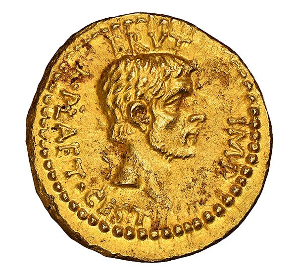 Close-up of an ancient gold Roman coin featuring the profile of a Roman emperor, with detailed engraving of the face and inscription around the edge. The coin's historical design is visible against a clean white background.
