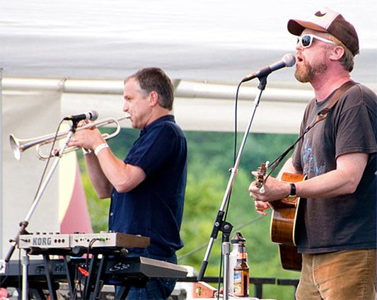 Two musicians performing on stage, one playing the trumpet and the other singing and playing the guitar, with a keyboard in the background and a beer bottle on stage.