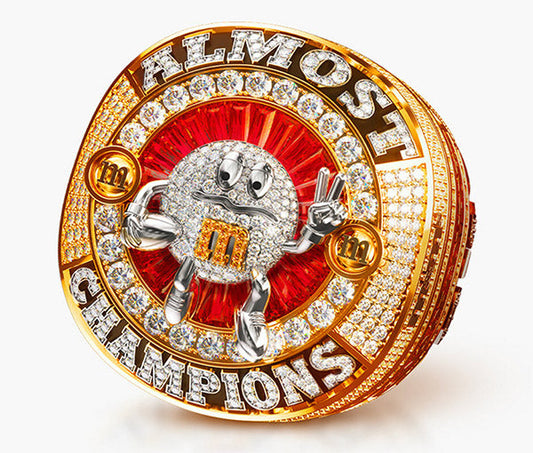 A unique, custom championship ring designed with the phrase "ALMOST CHAMPIONS" prominently displayed. The ring features the M&M character, which is adorned with diamonds and a bold red backdrop. 