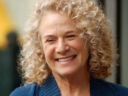 Portrait of singer-songwriter Carole King with curly hair, smiling and wearing a blue sweater.