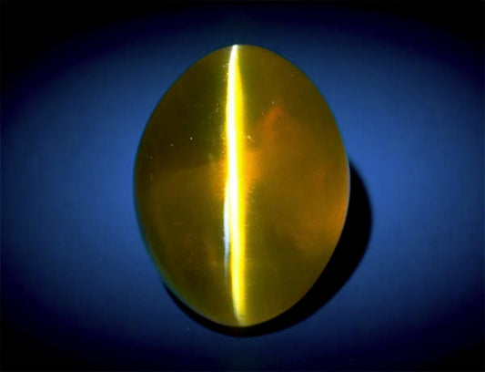 A close-up image of a large, yellow cat's eye gemstone with a prominent reflective line running vertically across the surface, displayed on a dark blue background.