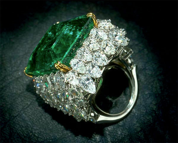 Close-up of a luxurious emerald ring with diamond accents, showcasing a large emerald stone surrounded by a halo of sparkling white diamonds on a silver band.
