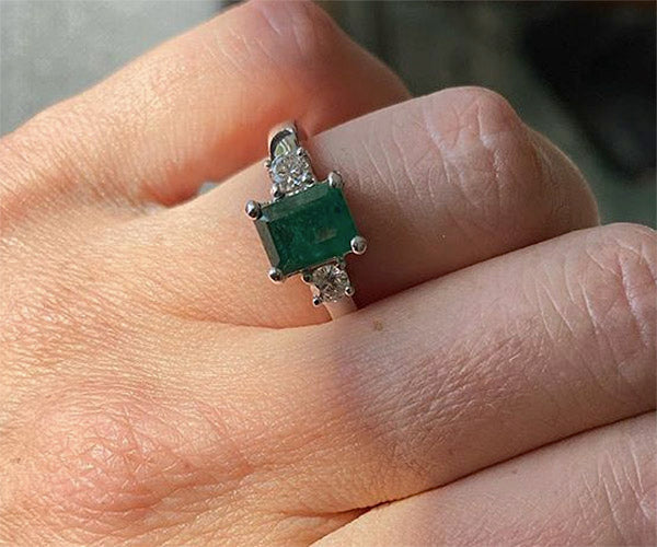 Close-up of a hand wearing a beautiful emerald engagement ring with a rectangular emerald center stone and diamond accents on either side.